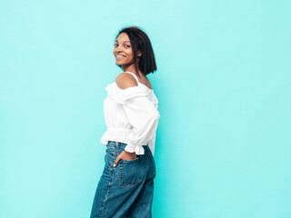 Portrait of young beautiful black woman. Smiling model dressed in summer jeans clothes. Sexy carefree female posing near blue wall in studio. Tanned and cheerful