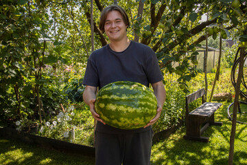 A smiling guy stands in a summer garden and holds a large ripe watermelon in his hands.