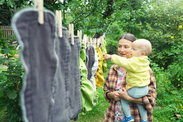 Mother with little baby hang reusable diapers to dry on clothesline in the garden. Modern eco...