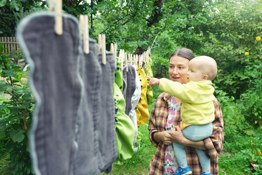 Mother with little baby hang reusable diapers to dry on clothesline in the garden. Modern eco friendly cloth nappy for infant child hygiene. Sustainable lifestyle, zero waste concept.