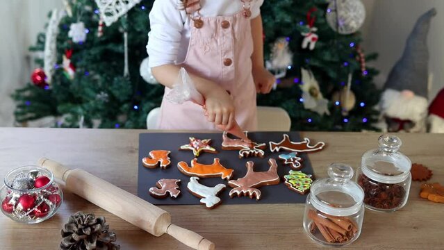Happy little girl coloring Christmas gingerbread cookies at home in slow motion. Christmas decoration in the kitchen. Fir tree with fairy lights. The concept of the New Year and Christmas. Holiday atm