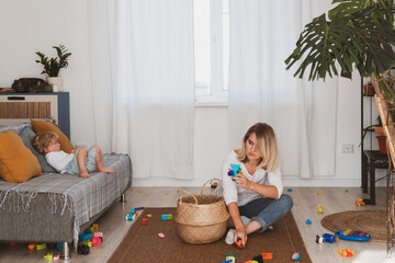 Young woman, housewife puts toys in the basket with her little son at home