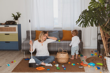 Young woman, housewife cleans up toys with broom together with her little son at home