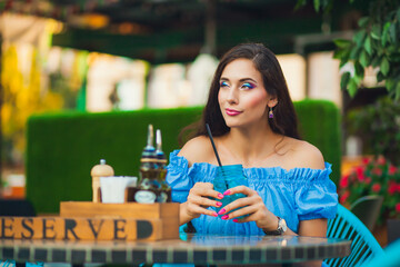 Young brunette woman with long hair in blue crop top is sitting at table in cafe and drinking from jar