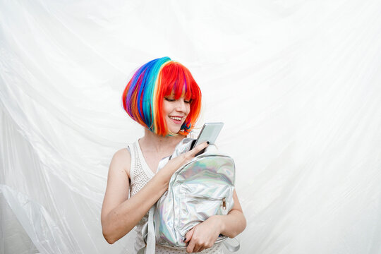 Smiling woman using smart phone with backpack in front of plastic backdrop