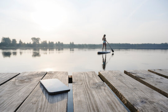 Mobile phone on jetty with woman doing standup paddleboarding in background at sunset