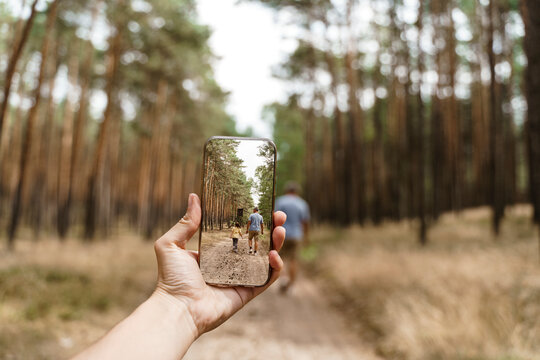 Woman With Smart Phone Taking Photo Of Man And Girl Walking In Forest