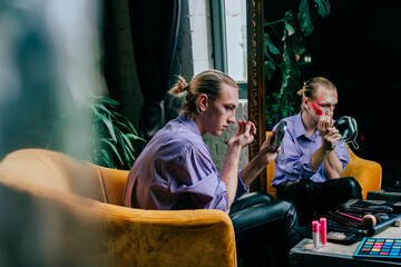 Man applying make-up sitting on chair by mirror
