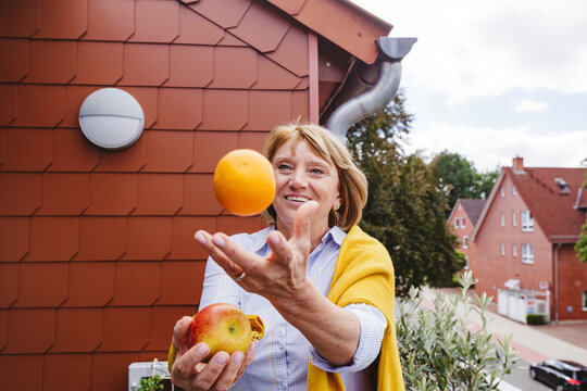 Happy senior woman juggling with orange and apple fruit in balcony
