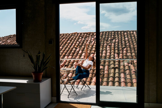 Woman with hand raised relaxing at balcony seen through window