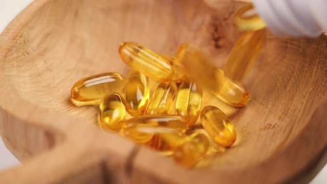 Health Supplement. Yellow Omega 3 capsules falling into a cracked wooden bowl from a white pharmaceutical plastic bottle. Rustic style view. Slow motion