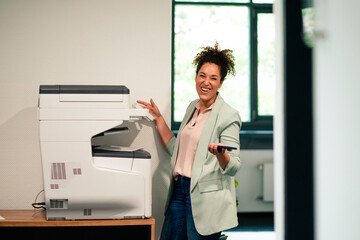 Cheerful businesswoman standing by computer printer at workplace