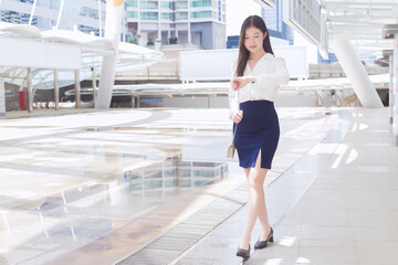 Young business Asian woman is walking to go to the office or workplace which she look at wrist watch in the big city with business buildings as a background.