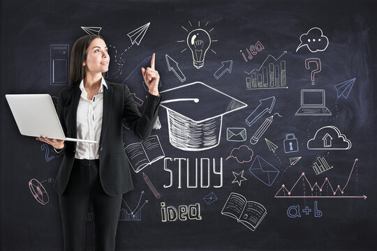 Attractive young european businesswoman with laptop pointing up at creative education sketch on chalkboard wall background. Freehand drawing of school items. Back to School concept.