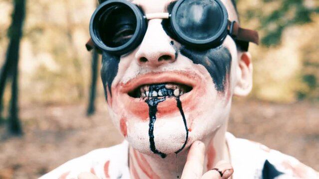 Portrait is close-up of face of zombie with traces of blood and black fluid flowing from the mouth. Man with makeup with aviation glasses with broken glass looks into camera and grimaces.
