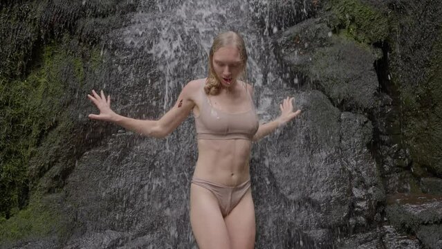 Blonde girl coming out of a waterfall in a wet swimsuit. Women's legs close-up, go to the stones, slow motion