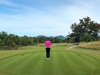 Obraz premium A rear view of lady stands focus aiming to the target on red tee off in the beautiful golf course with trees mountain and blue cloud sky in the background