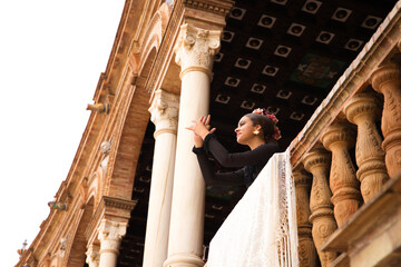 A beautiful teenage flamenco dancer with brown hair stands on a balcony between two white marble columns and performs flamenco dance postures and claps her hands artfully. - obrazy, fototapety, plakaty