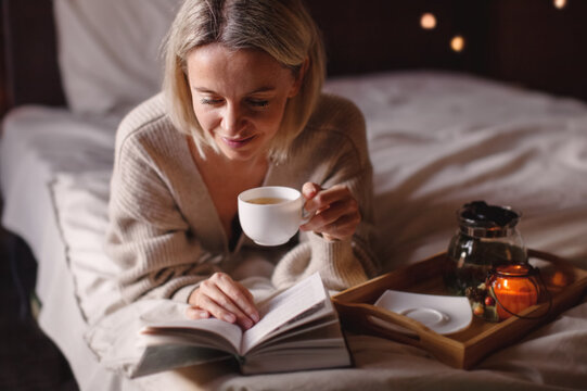 Relaxed middle aged woman sitting on bed reading book and drinking tea