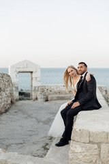 Portrait of happy couple sitting on coast of Adriatic sea on ancient stones. Young bride putting arm around grooms neck.