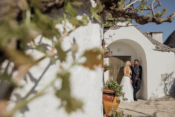 Portrait of wedding couple standing under arch of old white stone dwelling trullo with conical...