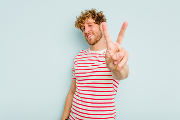Young caucasian man isolated on blue background joyful and carefree showing a peace symbol with...