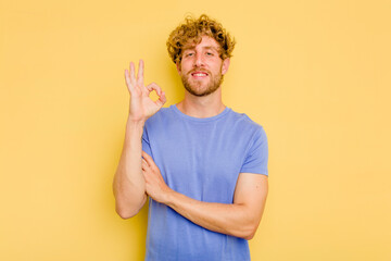 Young caucasian man isolated on yellow background winks an eye and holds an okay gesture with hand.