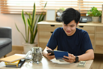 Satisfied asian man using smart phone for making online payments, managing expenses finances in living room