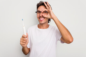 Young hispanic man holding electric toothbrush isolated on white background excited keeping ok gesture on eye.