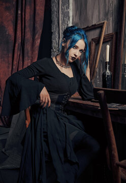 Indoors portrait of lovely goth girl in black dress. Blue-haired gothic lady sitting on chair at table. Vintage look