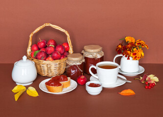 Tasty healthy homemade apple jam for breakfast. Wicker basket with small red apples, jars of fresh jam and cup of tea next to fallen leaves on brown table. Beautiful autumn still life