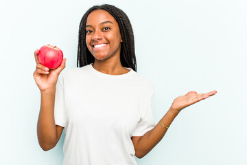 Young African American woman holding an apple isolated on blue background showing a copy space on a palm and holding another hand on waist.