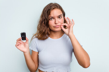 Young caucasian woman holding car keys isolated on blue background with fingers on lips keeping a secret.