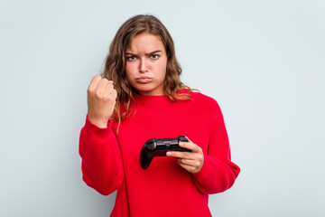 Young caucasian gamer woman holding a game controller isolated on blue background showing fist to camera, aggressive facial expression.