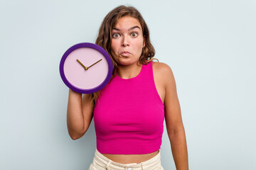 Young caucasian woman holding a clock isolated on blue background shrugs shoulders and open eyes confused.