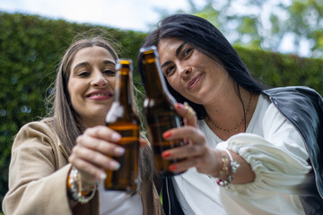 Two women friends sitting on the grass toasting with bottles of beer. Lifestyle of young friends in hats sitting on the grass toasting with wine