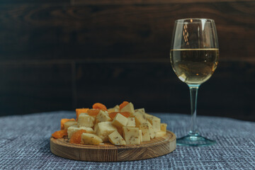 A glass of white wine and a slice of various types of cheese. Dark background. Snack for alcohol