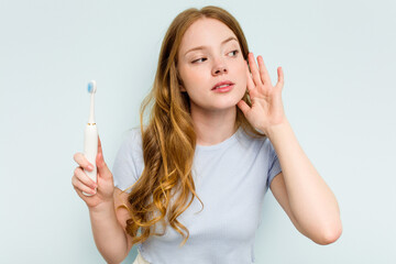 Young caucasian woman holding electric toothbrush isolated on blue background trying to listening a gossip.