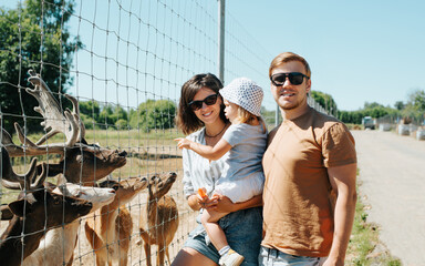 Happy beautiful family with baby at zoo farm outdoors. Smiling mom, dad with daughter in arms standing near animal fallow deer and looking at camera