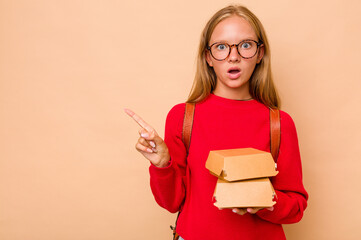 Little caucasian student girl holding burgers isolated on beige background pointing to the side