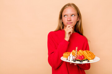 Little caucasian girl holding a waffles isolated on beige background looking sideways with doubtful and skeptical expression.