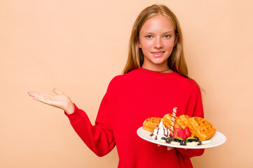 Little caucasian girl holding a waffles isolated on beige background showing a copy space on a palm and holding another hand on waist.