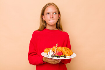 Little caucasian girl holding a waffles isolated on beige background confused, feels doubtful and unsure.