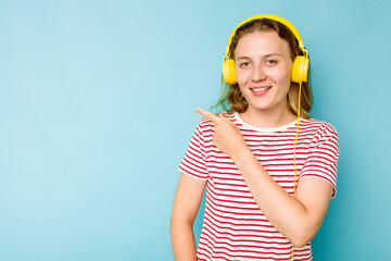 Young caucasian woman wearing headphones isolated on blue background smiling and pointing aside, showing something at blank space.