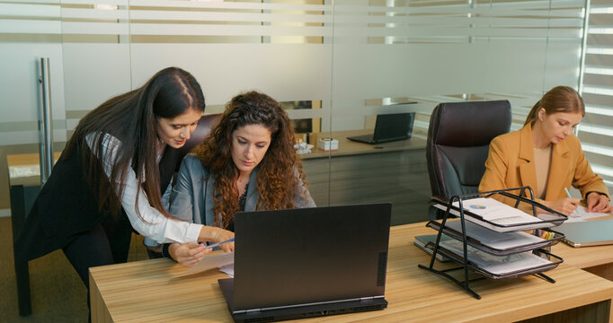 Three happy diverse women two having teamwork other looking into documents. Businesswomen solving challanges at workplace in office. Business concept.