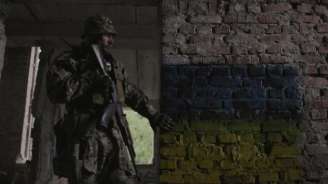 A Ukrainian military man with a weapon in his hands stands against the background of the flag of Ukraine. The flag of Ukraine is painted on a brick wall.