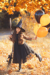 Happy little teen girl in black dress, leather jacket and witch hat with broom and balloons going...