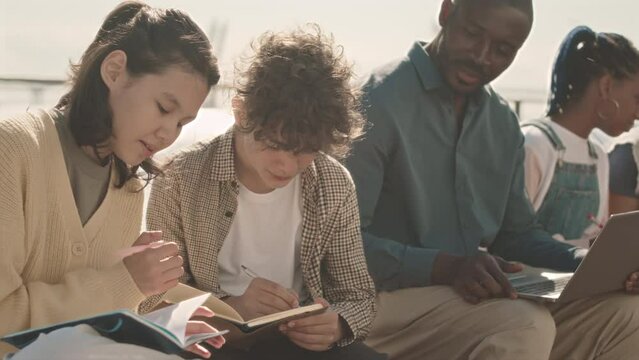 Medium slowmo of multiethnic schoolchildren taking notes in their copybooks while sitting on log at quay with their modern African American teacher, studying outdoors