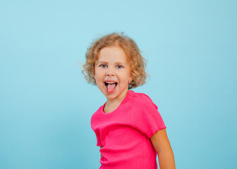 Portrait of happy blue-eyed little girl with curly hair looking, showing tongue, grimacing teasing on blue background.