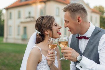 Side view of funny wedding couple standing outside near old building in park in summer, showing tongues to each other.